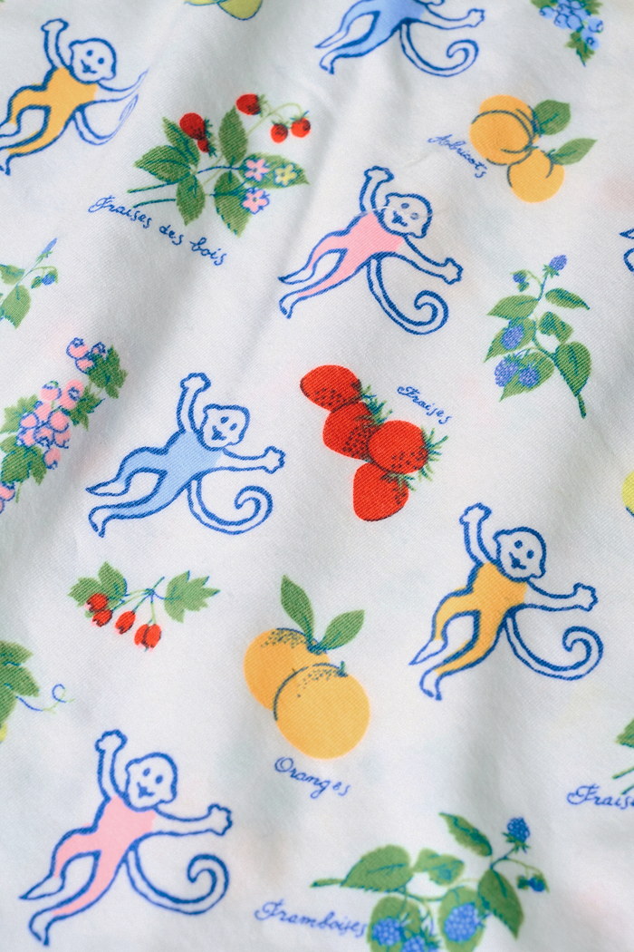 A person wearing a white sleeveless top and Roller Rabbit Monkey Fruits D'Ete Jones Shortie, featuring colorful prints of fruits, flowers, and playful cartoon figures; hands resting on the sides of the shorts.