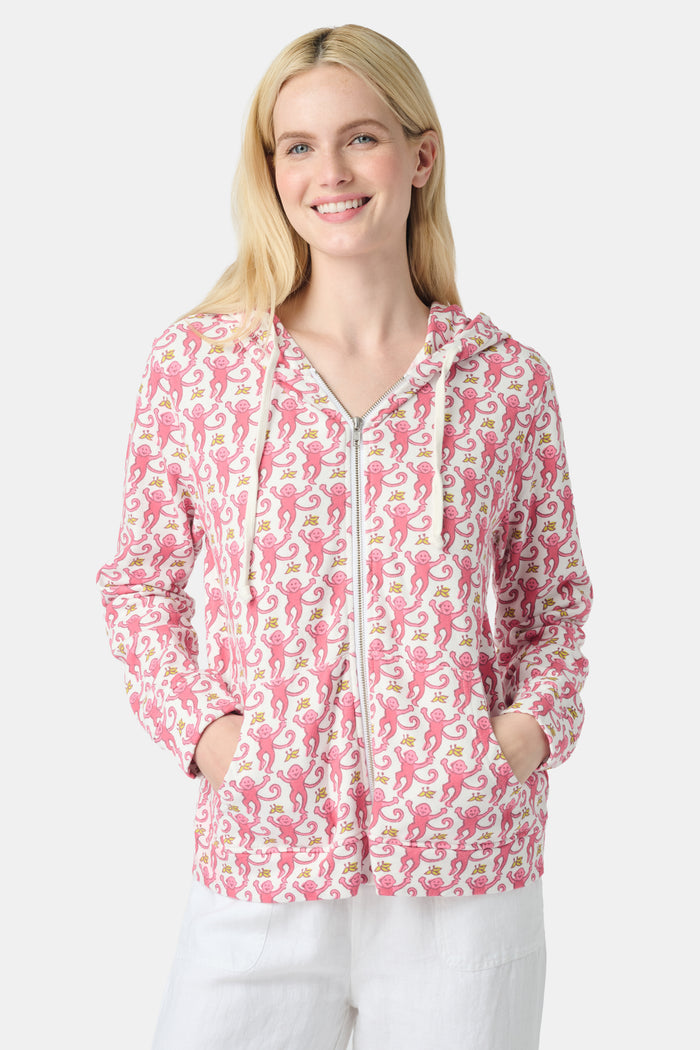 A smiling woman with long blonde hair wears the Roller Rabbit Monkey Harley Hoodie, a white zip-up French terry hoodie with pink seahorse pattern and white drawstrings, paired with white pants against a light background.