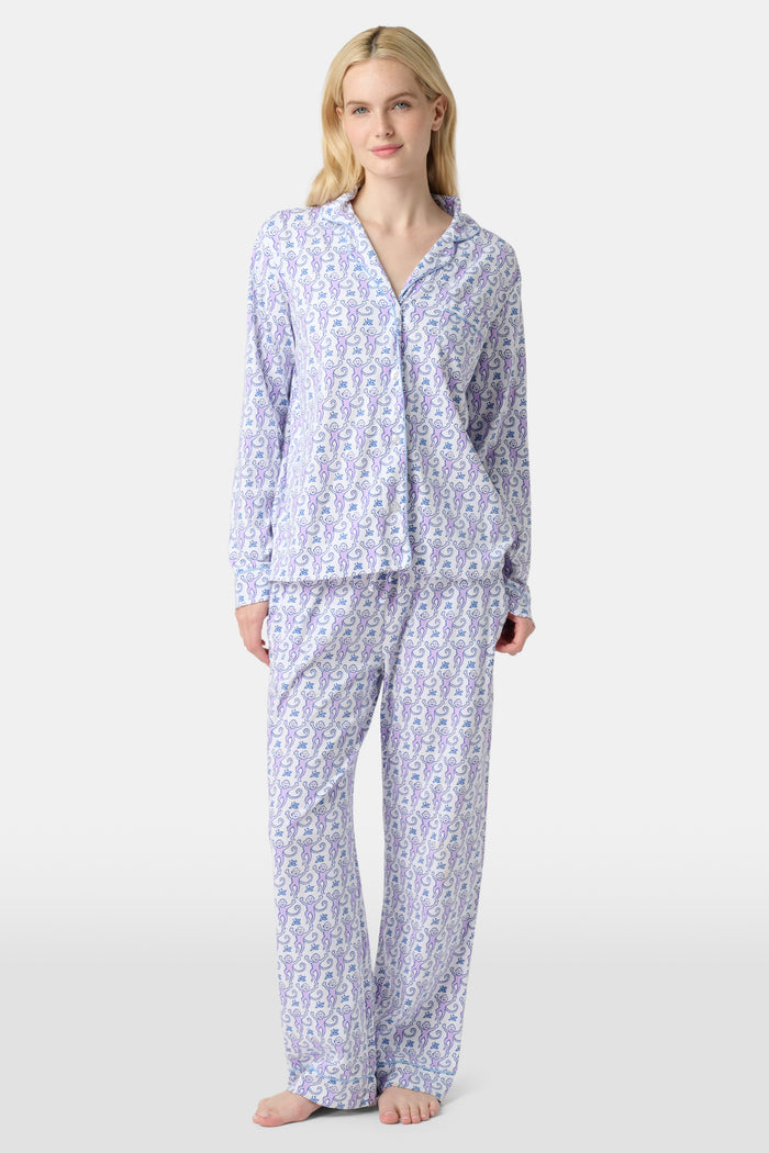 A woman stands barefoot against a plain, light background wearing the Roller Rabbit Monkey Long Sleeve Polo Pajamas, a women's set featuring a button-up top and loose-fitting pants in pima cotton with a light purple and white paisley print.