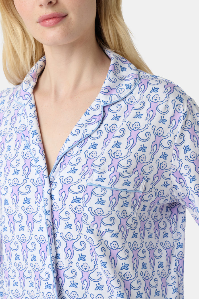 A woman stands barefoot against a plain, light background wearing the Roller Rabbit Monkey Long Sleeve Polo Pajamas, a women's set featuring a button-up top and loose-fitting pants in pima cotton with a light purple and white paisley print.