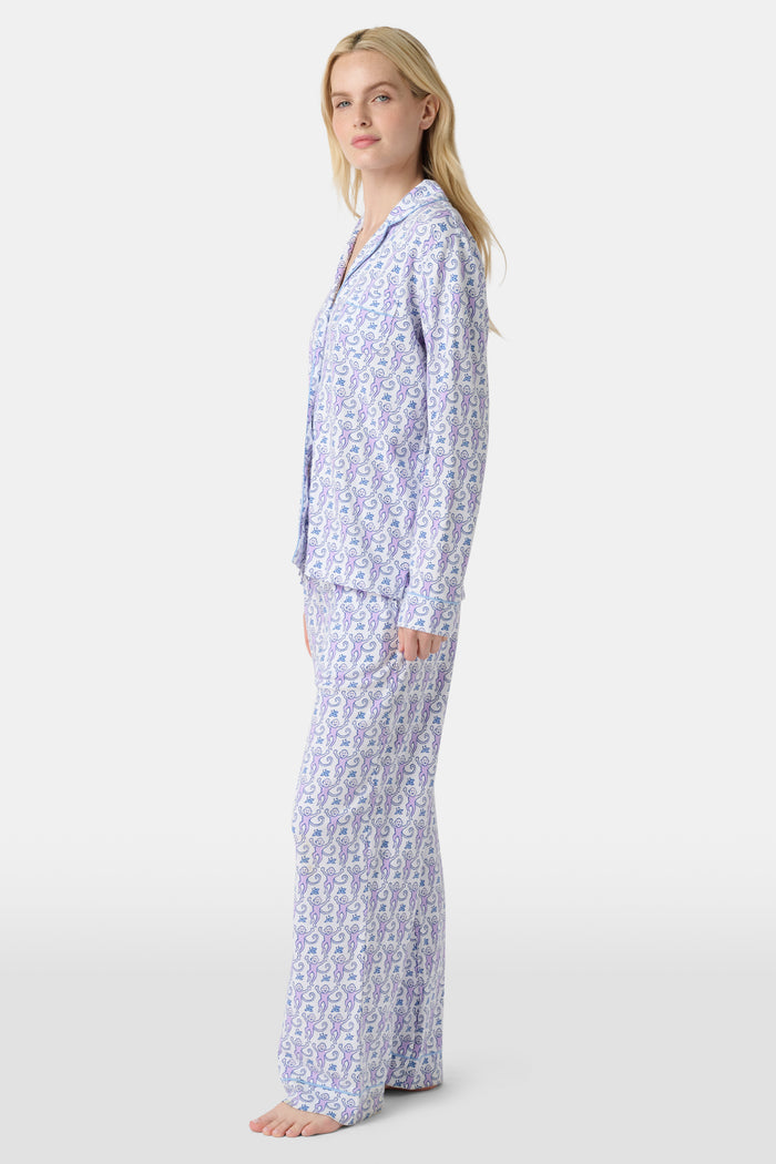 A woman stands barefoot against a plain, light background wearing the Roller Rabbit Monkey Long Sleeve Polo Pajamas, a women's set featuring a button-up top and loose-fitting pants in pima cotton with a light purple and white paisley print.