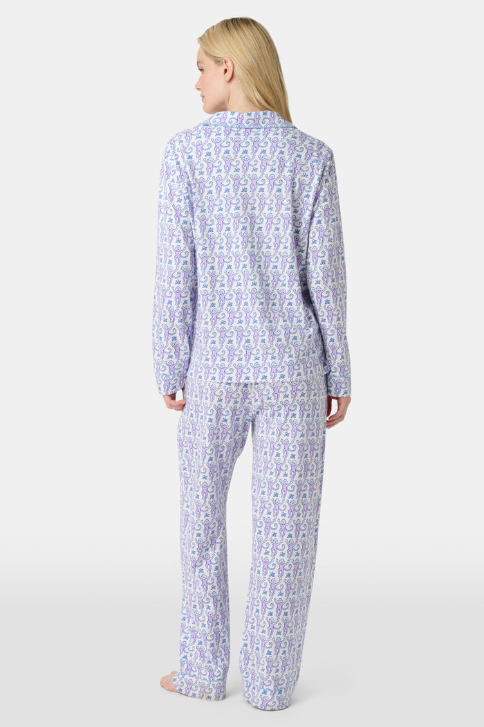 A woman stands barefoot against a plain, light background wearing the Roller Rabbit Monkey Long Sleeve Polo Pajamas, a women's set featuring a button-up top and loose-fitting pants in pima cotton with a light purple and white paisley print.