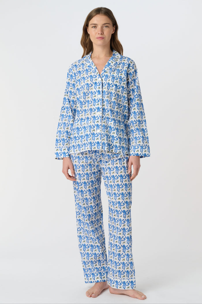 A person stands barefoot against a plain background, wearing the Roller Rabbit Monkey Loungewear set—a cotton pajama with a blue and white floral print, featuring a collared, button-up long sleeve top and loose-fitting pants.