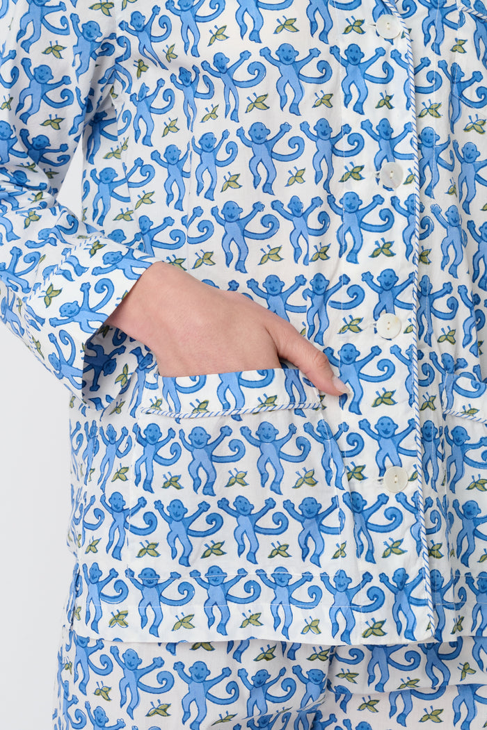 A person stands barefoot against a plain background, wearing the Roller Rabbit Monkey Loungewear set—a cotton pajama with a blue and white floral print, featuring a collared, button-up long sleeve top and loose-fitting pants.