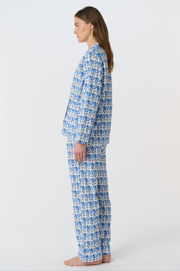 A person stands barefoot against a plain background, wearing the Roller Rabbit Monkey Loungewear set—a cotton pajama with a blue and white floral print, featuring a collared, button-up long sleeve top and loose-fitting pants.