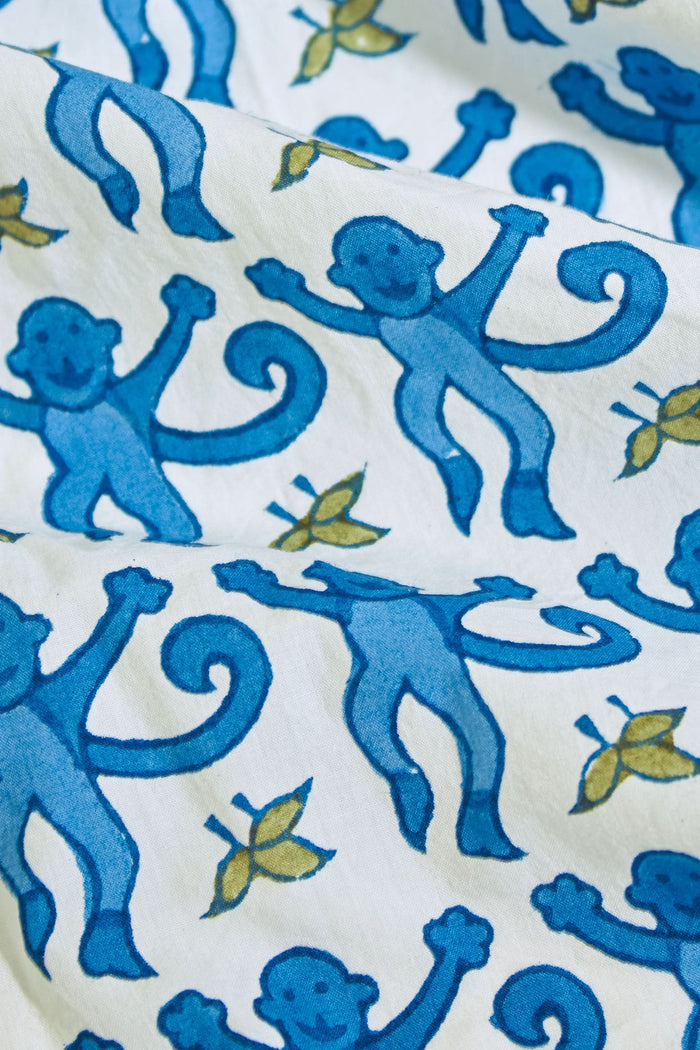 A person stands barefoot against a plain background, wearing the Roller Rabbit Monkey Loungewear set—a cotton pajama with a blue and white floral print, featuring a collared, button-up long sleeve top and loose-fitting pants.