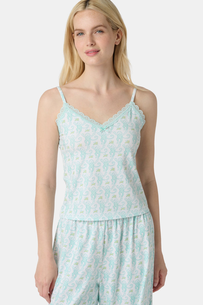 A woman with long blonde hair is wearing the Roller Rabbit Monkey Perry Cami pajama set. The light blue and white cotton-modal set features thin straps, a scalloped lace trim neckline, and matching loose pants. She stands, smiling slightly.