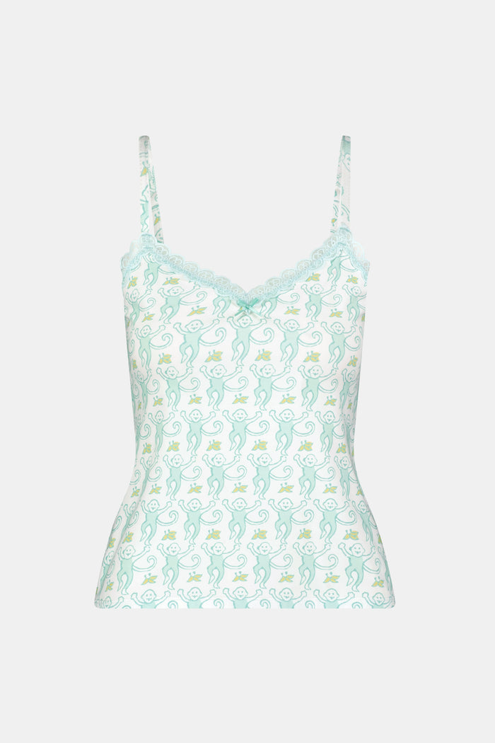 A woman with long blonde hair is wearing the Roller Rabbit Monkey Perry Cami pajama set. The light blue and white cotton-modal set features thin straps, a scalloped lace trim neckline, and matching loose pants. She stands, smiling slightly.