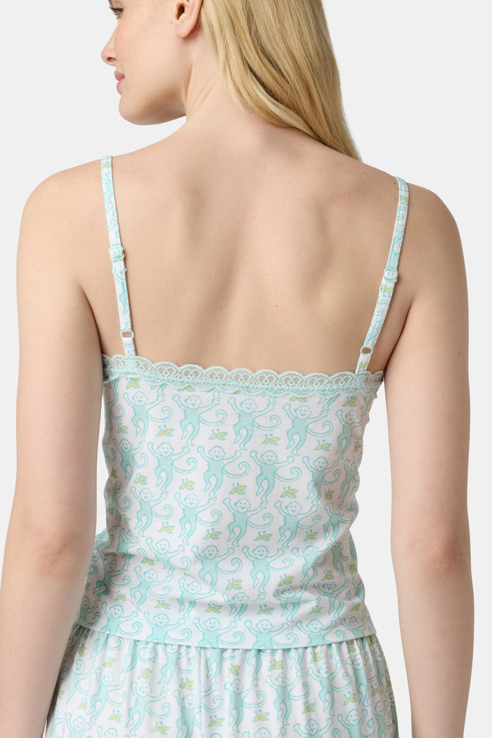 A woman with long blonde hair is wearing the Roller Rabbit Monkey Perry Cami pajama set. The light blue and white cotton-modal set features thin straps, a scalloped lace trim neckline, and matching loose pants. She stands, smiling slightly.
