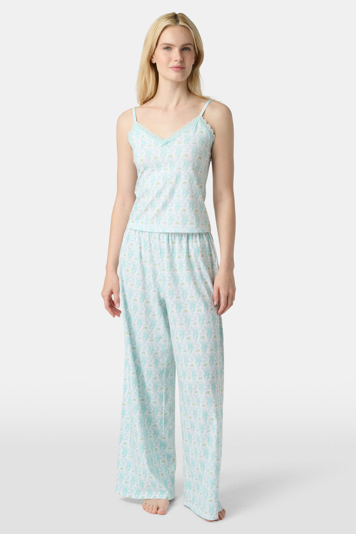 A woman with long blonde hair is wearing the Roller Rabbit Monkey Perry Cami pajama set. The light blue and white cotton-modal set features thin straps, a scalloped lace trim neckline, and matching loose pants. She stands, smiling slightly.