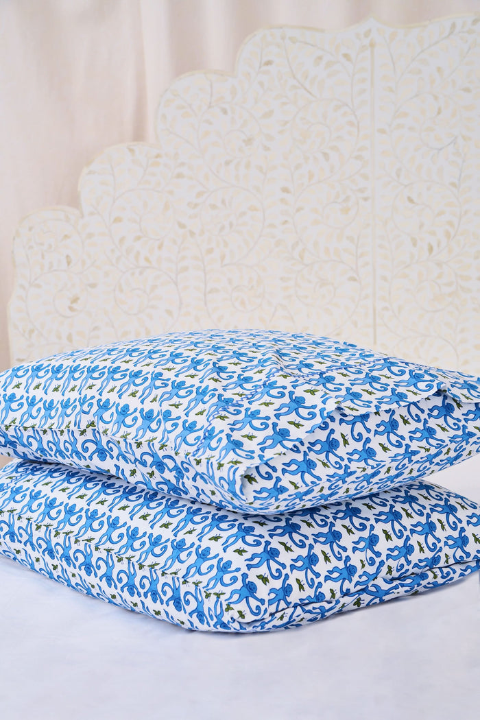 Two Monkey Pillowcase Set pillows by Roller Rabbit, featuring a blue and green floral pattern and 100 cotton pillowcases, are stacked on a white surface with a cream decorative screen in the background.