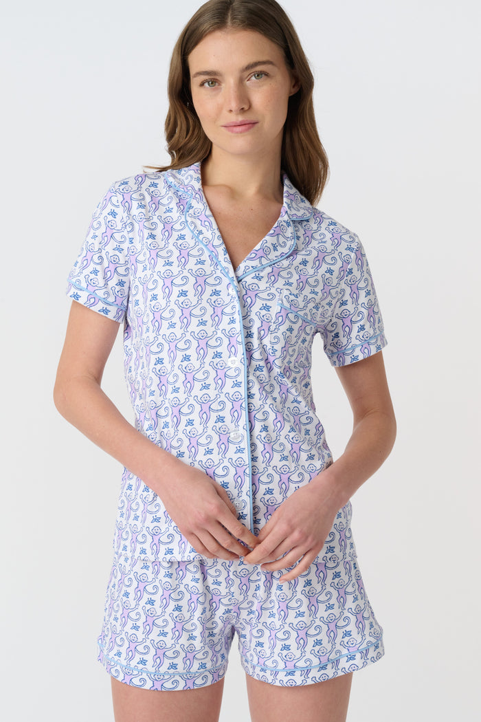 A woman with long brown hair wears Roller Rabbit's Monkey Polo Pajamas, a short-sleeve button-up Pima cotton romper featuring a blue and purple floral pattern, standing against a plain white background.