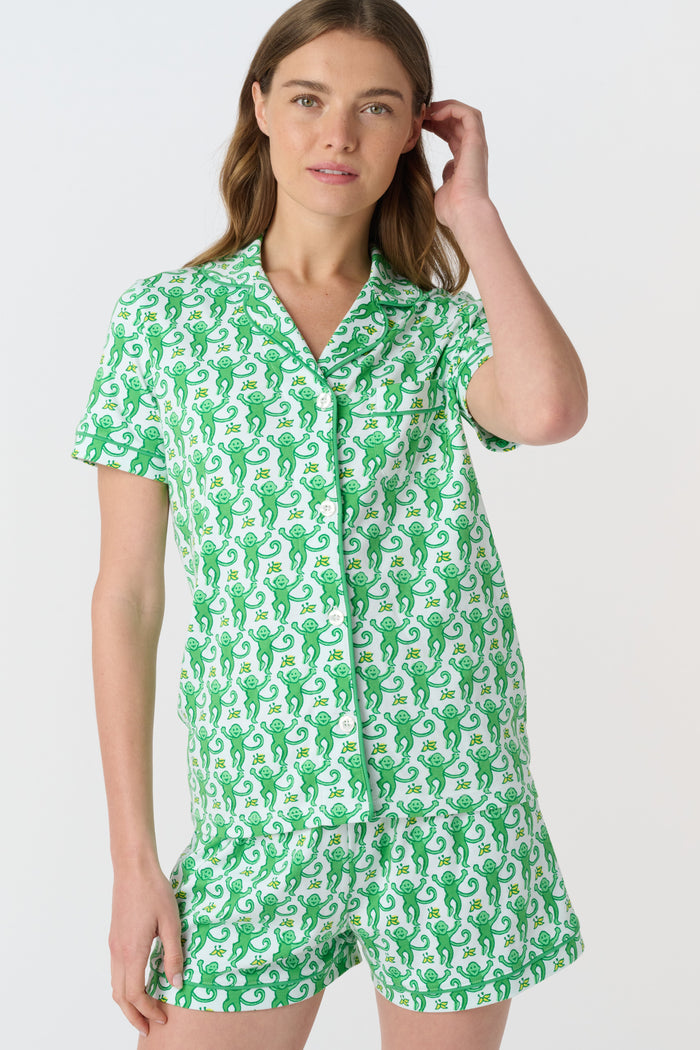 A woman wears Roller Rabbit's Monkey Polo Pajamas, a matching green and white monkey-patterned set with a button-up top and shorts. She touches her hair and smiles softly, looking slightly to the side in her stylish sleepwear.