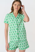 A woman wears Roller Rabbit's Monkey Polo Pajamas, a matching green and white monkey-patterned set with a button-up top and shorts. She touches her hair and smiles softly, looking slightly to the side in her stylish sleepwear.