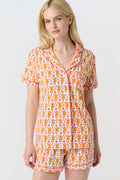 A woman with long blonde hair wears the Monkey Polo Pajamas by Roller Rabbit, a soft Pima cotton set featuring a playful orange and pink cat print, as she smiles gently at the camera against a plain white background.