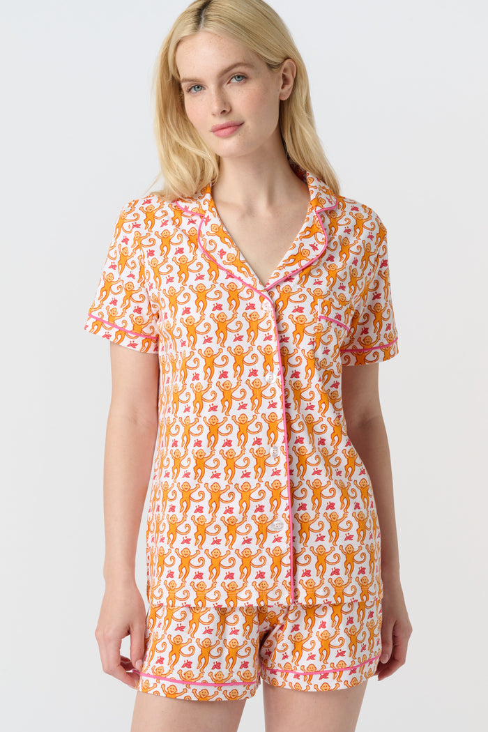 A woman with long blonde hair wears the Monkey Polo Pajamas by Roller Rabbit, a soft Pima cotton set featuring a playful orange and pink cat print, as she smiles gently at the camera against a plain white background.