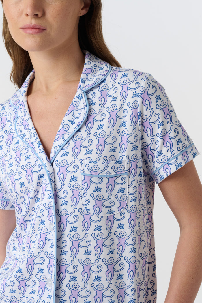 A woman with long brown hair wears Roller Rabbit's Monkey Polo Pajamas, a short-sleeve button-up Pima cotton romper featuring a blue and purple floral pattern, standing against a plain white background.