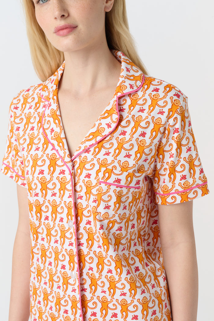 A woman with long blonde hair wears the Monkey Polo Pajamas by Roller Rabbit, a soft Pima cotton set featuring a playful orange and pink cat print, as she smiles gently at the camera against a plain white background.