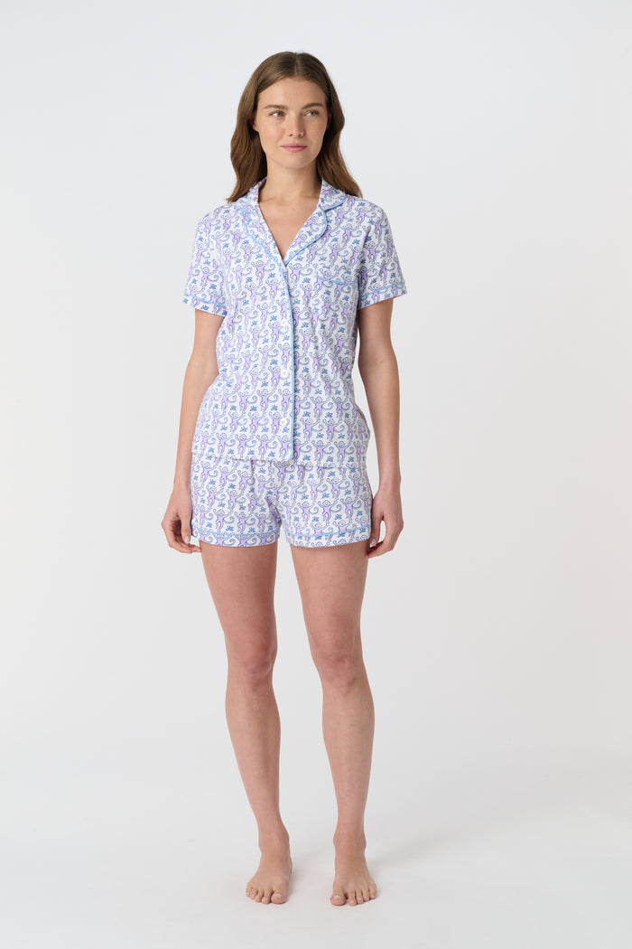 A woman with long brown hair wears Roller Rabbit's Monkey Polo Pajamas, a short-sleeve button-up Pima cotton romper featuring a blue and purple floral pattern, standing against a plain white background.