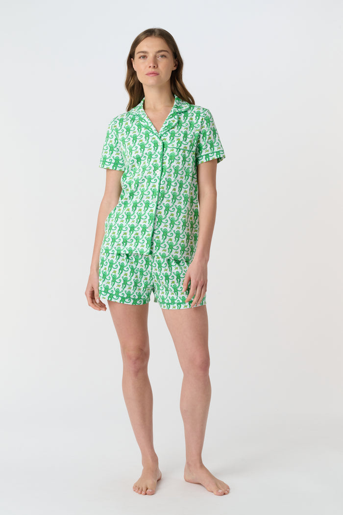 A woman wears Roller Rabbit's Monkey Polo Pajamas, a matching green and white monkey-patterned set with a button-up top and shorts. She touches her hair and smiles softly, looking slightly to the side in her stylish sleepwear.