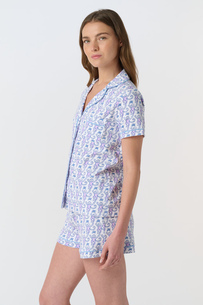 A woman with long brown hair wears Roller Rabbit's Monkey Polo Pajamas, a short-sleeve button-up Pima cotton romper featuring a blue and purple floral pattern, standing against a plain white background.