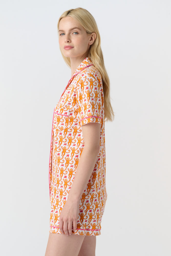 A woman with long blonde hair wears the Monkey Polo Pajamas by Roller Rabbit, a soft Pima cotton set featuring a playful orange and pink cat print, as she smiles gently at the camera against a plain white background.