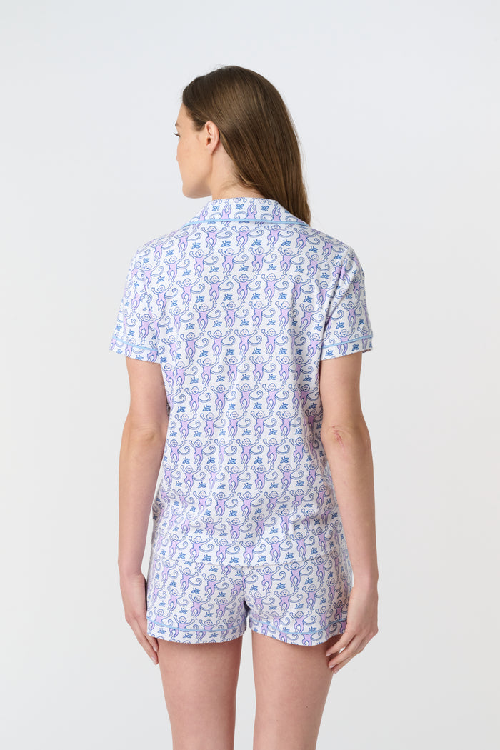 A woman with long brown hair wears Roller Rabbit's Monkey Polo Pajamas, a short-sleeve button-up Pima cotton romper featuring a blue and purple floral pattern, standing against a plain white background.