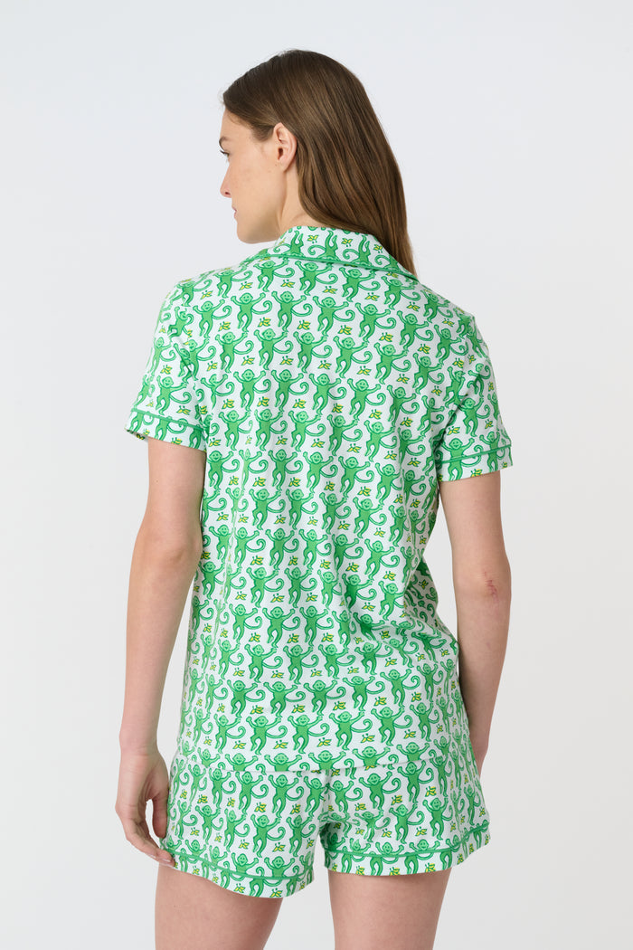 A woman wears Roller Rabbit's Monkey Polo Pajamas, a matching green and white monkey-patterned set with a button-up top and shorts. She touches her hair and smiles softly, looking slightly to the side in her stylish sleepwear.