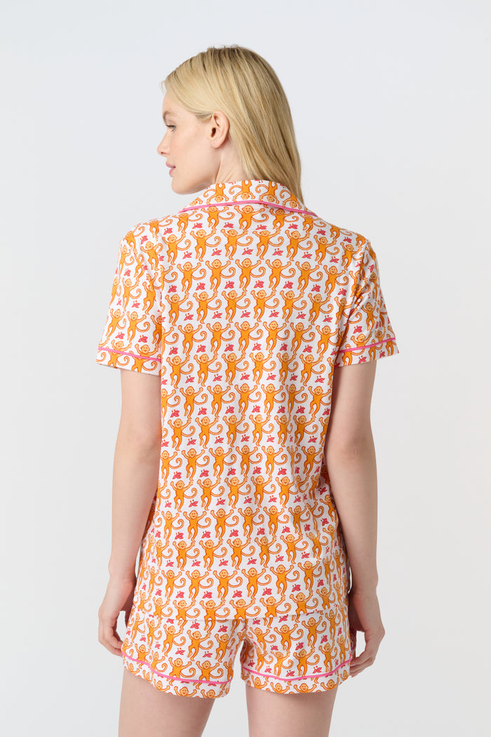 A woman with long blonde hair wears the Monkey Polo Pajamas by Roller Rabbit, a soft Pima cotton set featuring a playful orange and pink cat print, as she smiles gently at the camera against a plain white background.