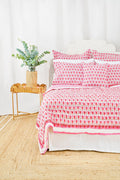 A bed with the Roller Rabbit Monkey Quilt in white and pink cotton and pillows stands by a woven side table with a vase of green eucalyptus, all on a natural fiber rug against a white background.