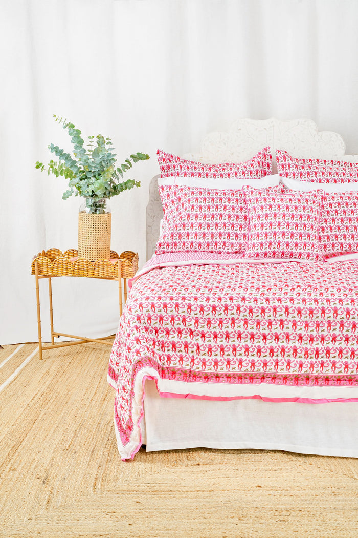 A bed with the Roller Rabbit Monkey Quilt in white and pink cotton and pillows stands by a woven side table with a vase of green eucalyptus, all on a natural fiber rug against a white background.