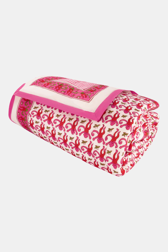 A bed with the Roller Rabbit Monkey Quilt in white and pink cotton and pillows stands by a woven side table with a vase of green eucalyptus, all on a natural fiber rug against a white background.