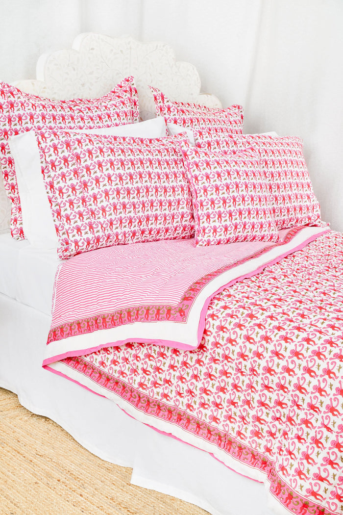 A bed with the Roller Rabbit Monkey Quilt in white and pink cotton and pillows stands by a woven side table with a vase of green eucalyptus, all on a natural fiber rug against a white background.