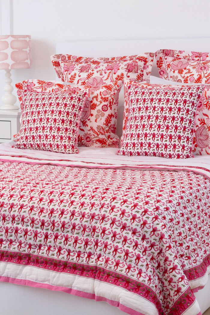 A bed with the Roller Rabbit Monkey Quilt in white and pink cotton and pillows stands by a woven side table with a vase of green eucalyptus, all on a natural fiber rug against a white background.