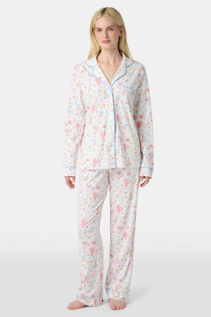 A woman stands barefoot in Roller Rabbit’s Monkey Said Yes Long Sleeve Polo Pajamas—white with a pink and blue floral pattern and blue trim—against a plain white background, perfect for bridal getting-ready moments.