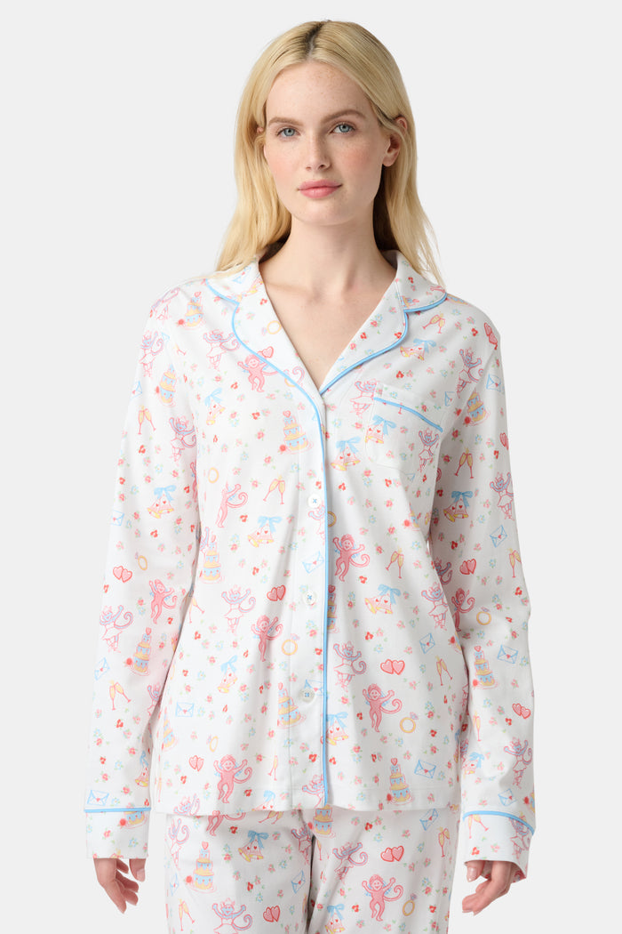A woman stands barefoot in Roller Rabbit’s Monkey Said Yes Long Sleeve Polo Pajamas—white with a pink and blue floral pattern and blue trim—against a plain white background, perfect for bridal getting-ready moments.