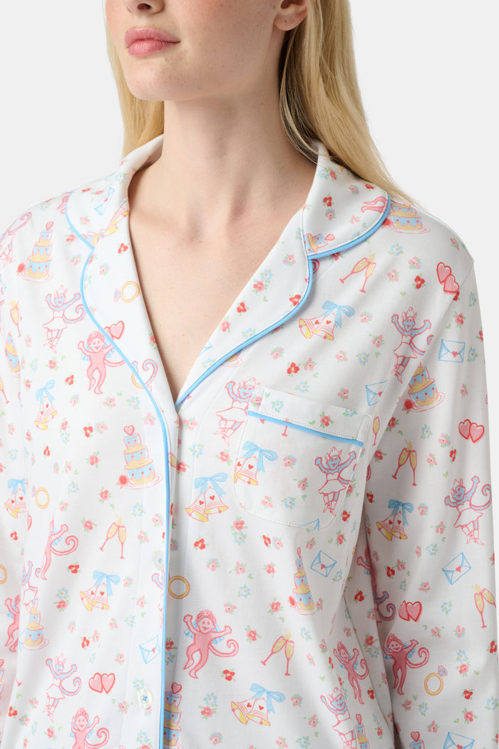 A woman stands barefoot in Roller Rabbit’s Monkey Said Yes Long Sleeve Polo Pajamas—white with a pink and blue floral pattern and blue trim—against a plain white background, perfect for bridal getting-ready moments.