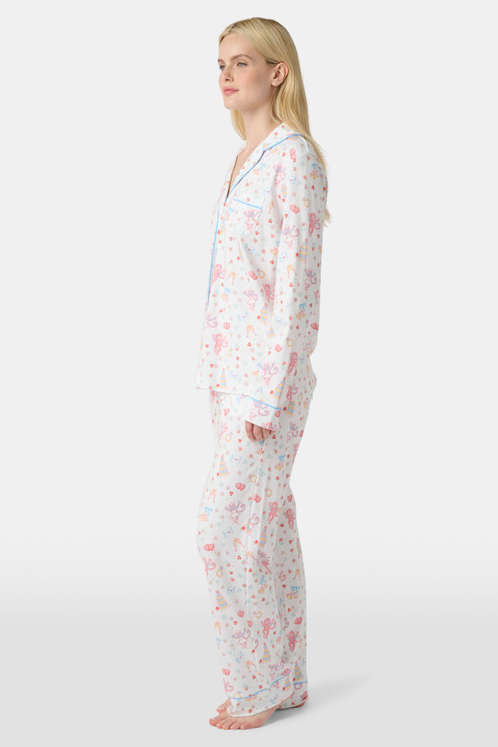 A woman stands barefoot in Roller Rabbit’s Monkey Said Yes Long Sleeve Polo Pajamas—white with a pink and blue floral pattern and blue trim—against a plain white background, perfect for bridal getting-ready moments.