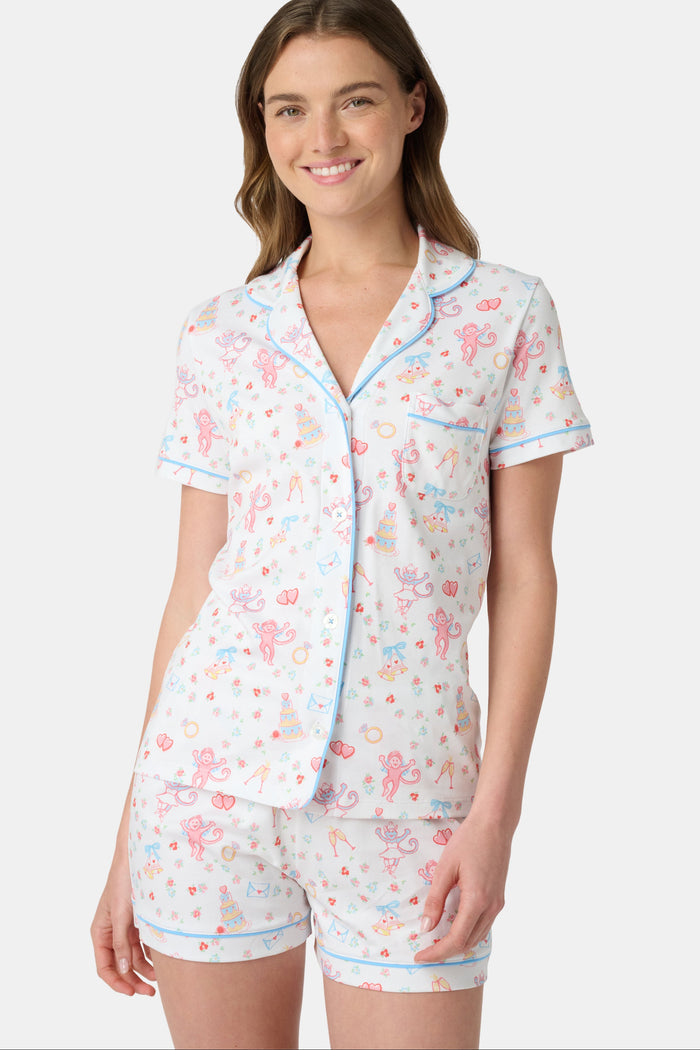 A woman smiles while wearing Roller Rabbit’s Monkey Said Yes Polo Pajamas—a short-sleeved, button-up top and matching shorts with a colorful, playful print featuring whimsical illustrations.