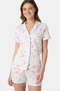 A woman smiles while wearing Roller Rabbit’s Monkey Said Yes Polo Pajamas—a short-sleeved, button-up top and matching shorts with a colorful, playful print featuring whimsical illustrations.