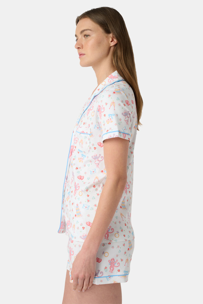 A woman smiles while wearing Roller Rabbit’s Monkey Said Yes Polo Pajamas—a short-sleeved, button-up top and matching shorts with a colorful, playful print featuring whimsical illustrations.