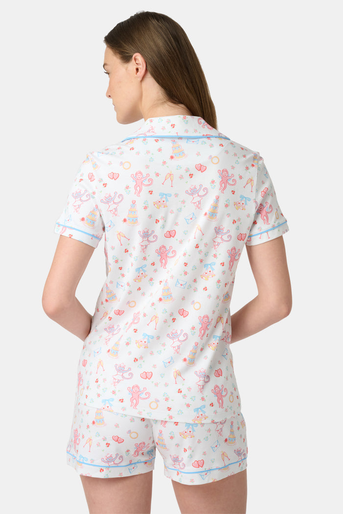 A woman smiles while wearing Roller Rabbit’s Monkey Said Yes Polo Pajamas—a short-sleeved, button-up top and matching shorts with a colorful, playful print featuring whimsical illustrations.