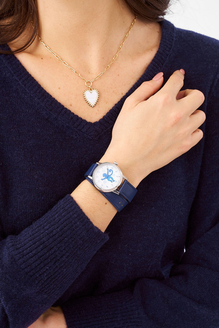 A woman wears a navy blue sweater, a gold heart-shaped pendant necklace, and the Monkey Watch by Roller Rabbit with an Italian leather strap. She crosses her arm over her chest, highlighting her stylish accessories.