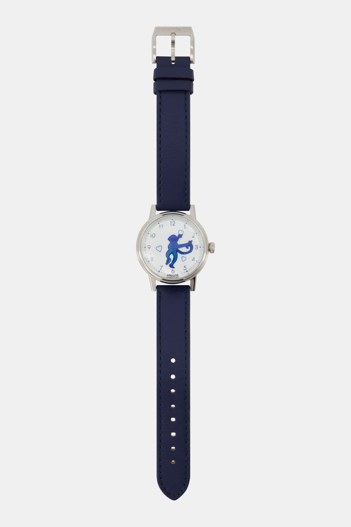 A woman wears a navy blue sweater, a gold heart-shaped pendant necklace, and the Monkey Watch by Roller Rabbit with an Italian leather strap. She crosses her arm over her chest, highlighting her stylish accessories.