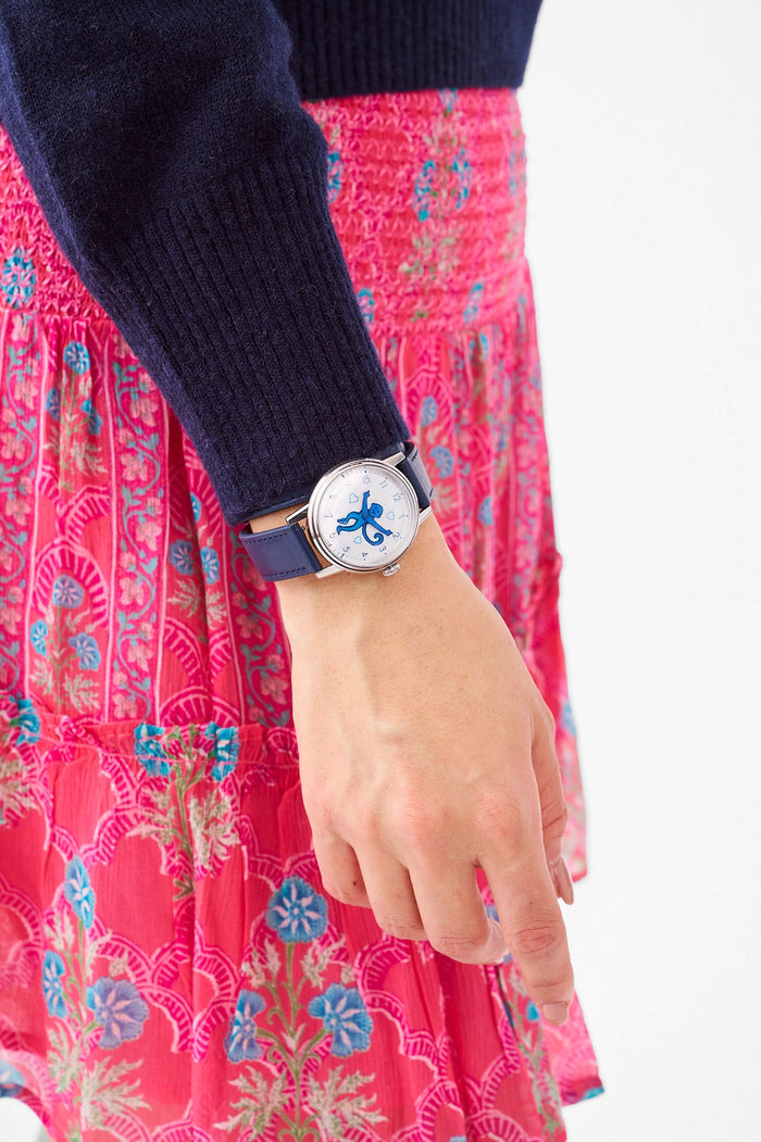 A woman wears a navy blue sweater, a gold heart-shaped pendant necklace, and the Monkey Watch by Roller Rabbit with an Italian leather strap. She crosses her arm over her chest, highlighting her stylish accessories.