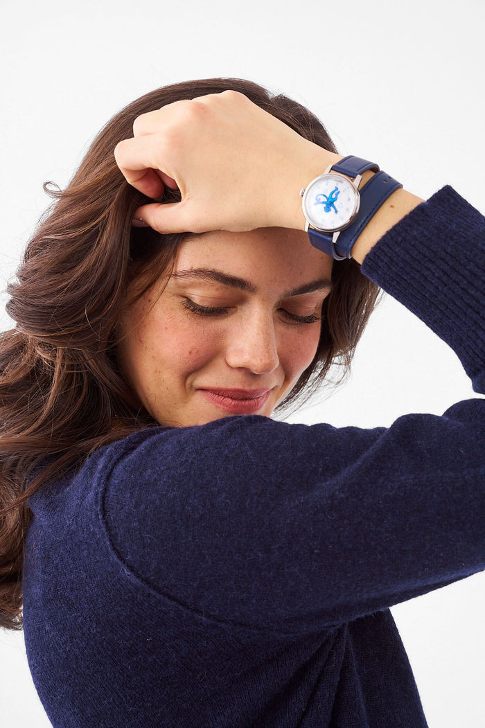 A woman wears a navy blue sweater, a gold heart-shaped pendant necklace, and the Monkey Watch by Roller Rabbit with an Italian leather strap. She crosses her arm over her chest, highlighting her stylish accessories.