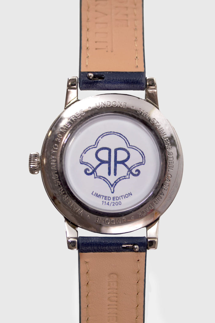 A woman wears a navy blue sweater, a gold heart-shaped pendant necklace, and the Monkey Watch by Roller Rabbit with an Italian leather strap. She crosses her arm over her chest, highlighting her stylish accessories.