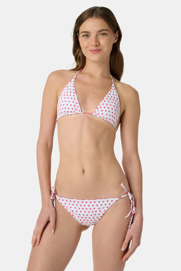 A woman wearing the Roller Rabbit Monkey/Hearts Serena Bikini Top and a straw hat smiles happily on a beach with the ocean in the background.