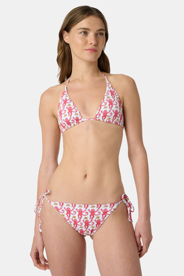 A woman wearing the Roller Rabbit Monkey/Hearts Serena Bikini Top and a straw hat smiles happily on a beach with the ocean in the background.