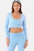 A woman stands facing forward against a plain light background, wearing the Roller Rabbit Morgan Wrap Top in sky blue, paired with high-waisted leggings and a matching headband, modeling stylish women’s activewear.
